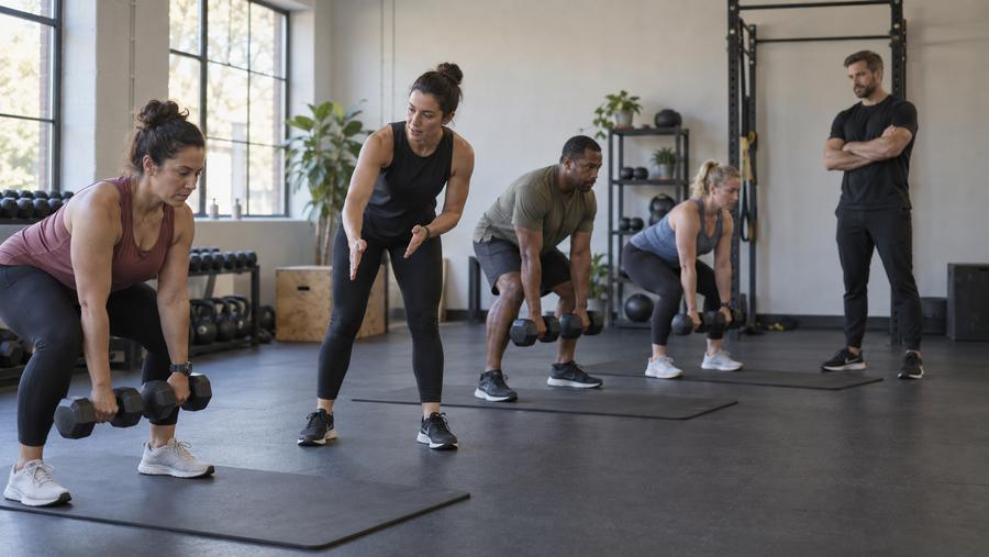 A head coach watches from the side as another coach cues members using dumbbells in a group strength class