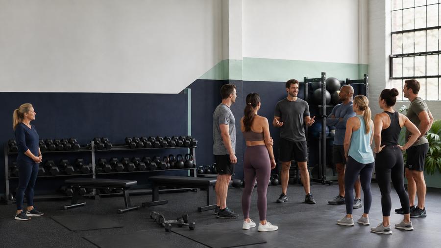A new coach welcoming group fitness members while a site manager observes in an unbranded functional training studio